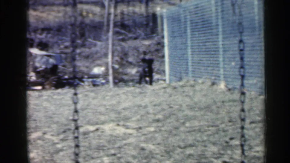 1959 Indiana 8mm frame showing a yard, chain-link fence, bare trees, a swing chain in the foreground, and a small dark animal near the fence line.