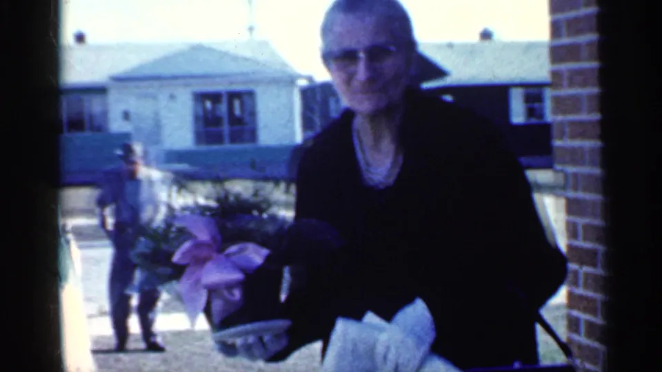 1959 Indiana 8mm frame showing a woman in glasses holding colorful flowers near a brick house exterior, with another person behind her.