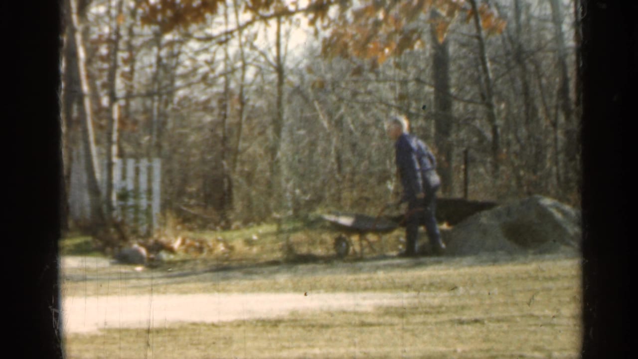 Man standing beside a wheelbarrow near a dirt pile in a grassy wooded yard.