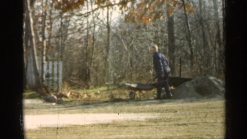 In 1959 Indiana home-movie footage, a man in a light hat stands by a wheelbarrow near a dirt pile, trees and a white fence.