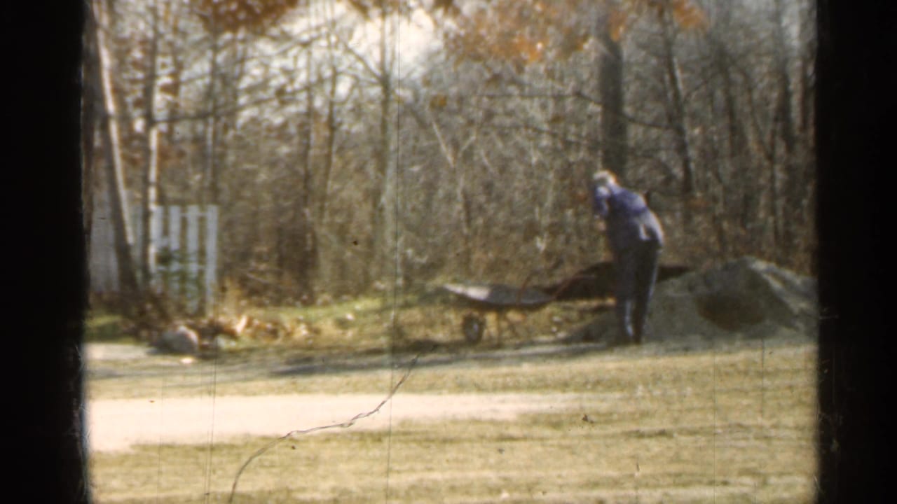 Man bends near a wheelbarrow and dirt pile in a sunlit yard.