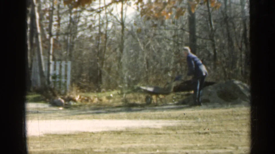 A man in dark clothing and a light hat handles a wheelbarrow beside a dirt pile in 1959 Indiana home-movie footage.