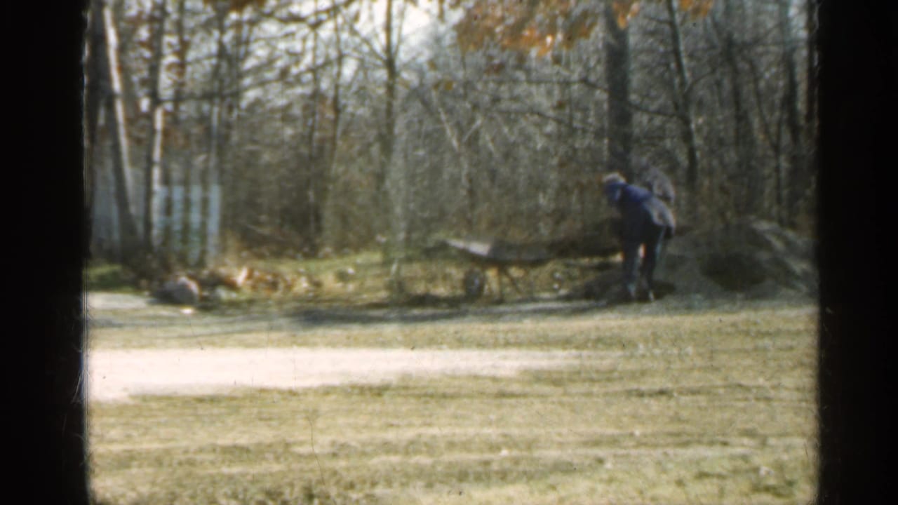 Man in a blue jacket works beside a wheelbarrow in a wooded backyard.