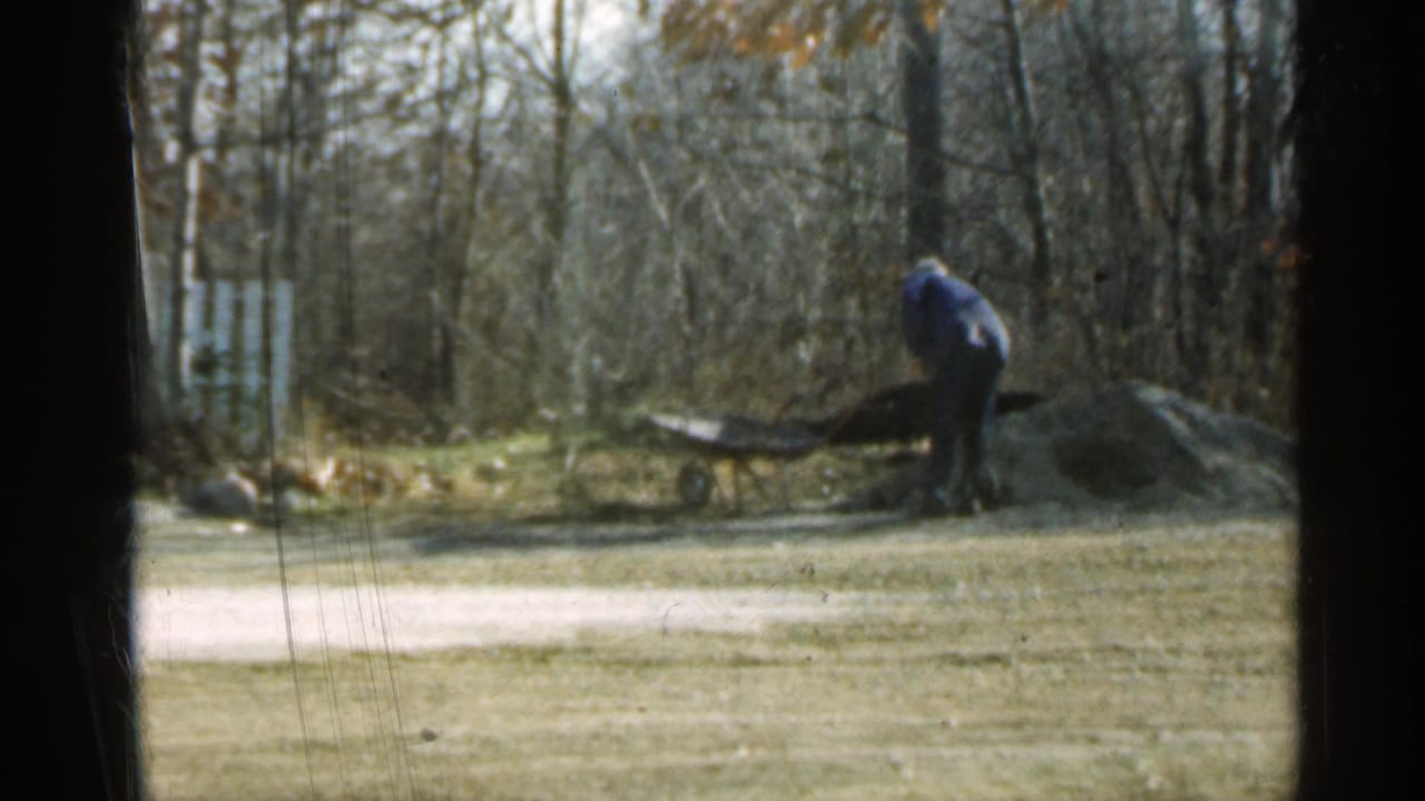 Man bends near a wheelbarrow beside a dirt pile in the yard.
