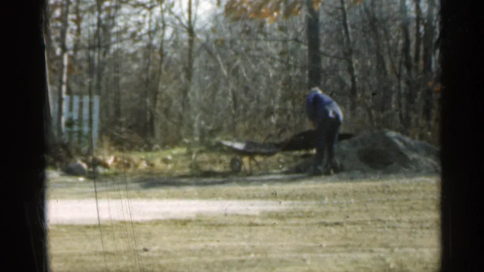 1959 Indiana frame with an adult man bending by a wheelbarrow near a dirt or mulch pile in a residential yard.