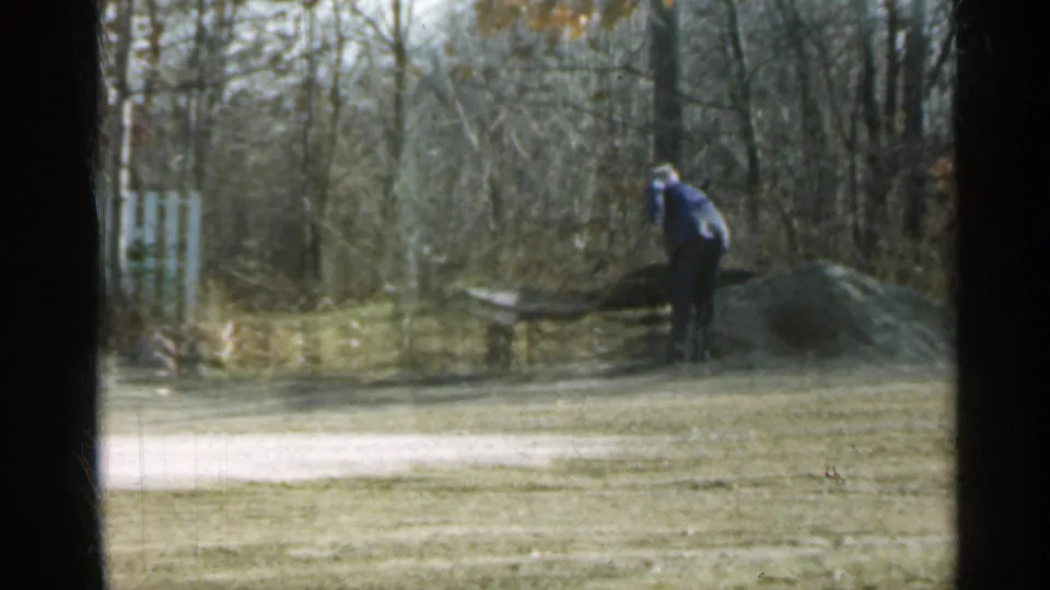 1959 Indiana home-movie view of a man handling a wheelbarrow beside a low pile, with lawn and leafless trees around him.