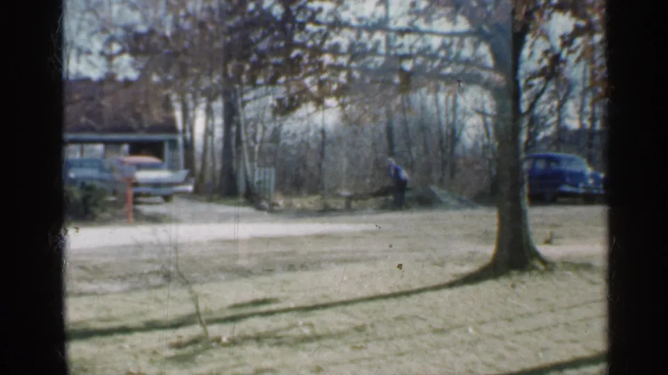 Wide 1959 Indiana home-movie frame showing the man working near a pile, with driveway, trees, and parked cars in the background.