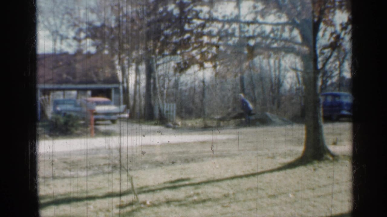 Indiana 1959 frame of a residential yard with two cars near a driveway, bare trees, a house, and a distant person working near brush.