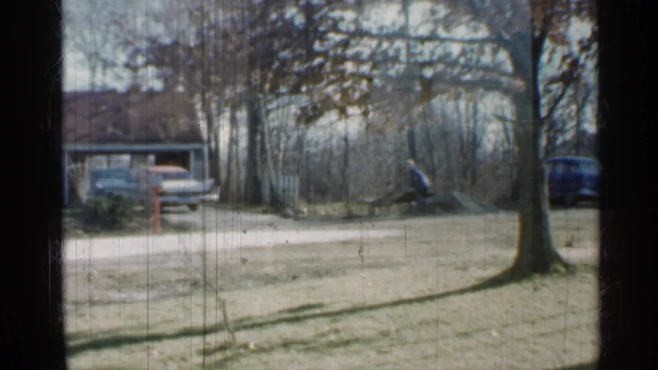 Indiana 1959 frame of a residential yard with two cars near a driveway, bare trees, a house, and a distant person working near brush.