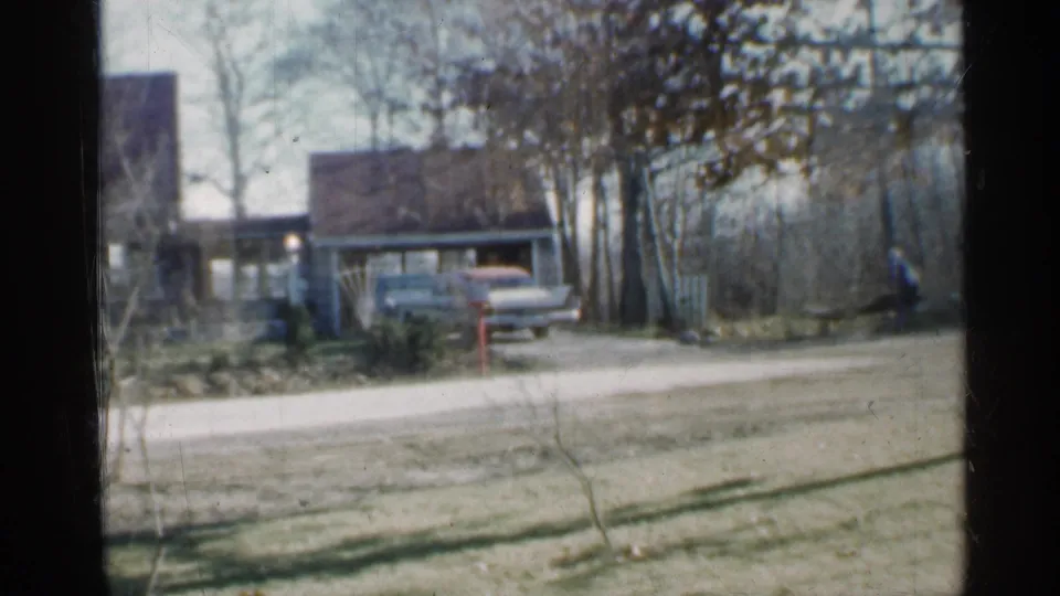 Indiana 1959 home-movie view from indoors toward a house and driveway with parked cars, leafless trees, lawn, and a distant person outside.