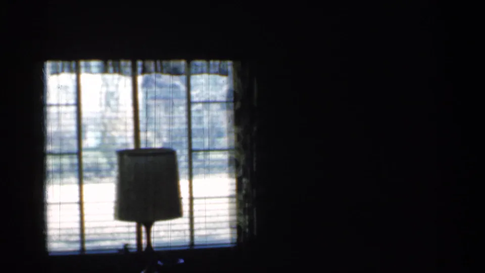 1959 Indiana home-movie frame showing a dim room with a lamp silhouetted against a bright window and blinds.