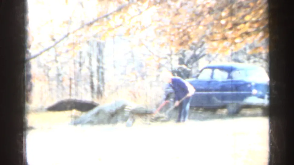 1959 Indiana yard scene with a man shoveling near piles of dirt and a blue mid-century car beneath autumn trees.