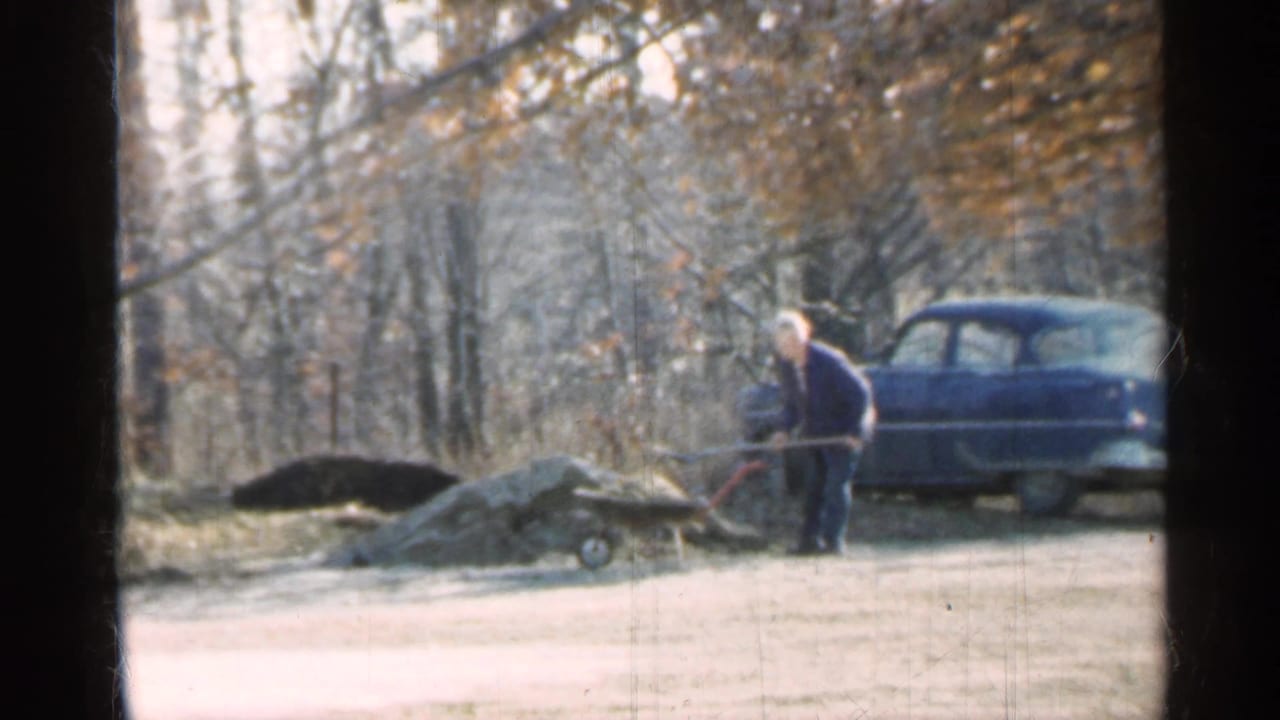 Man shoveling near a wheelbarrow and dirt piles with a blue sedan behind him.