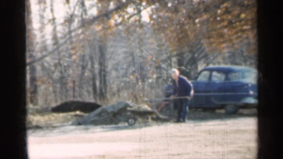 1959 Indiana 8mm frame showing a man shoveling dirt or rocks beside a wheelbarrow, with a blue sedan parked near bare trees.