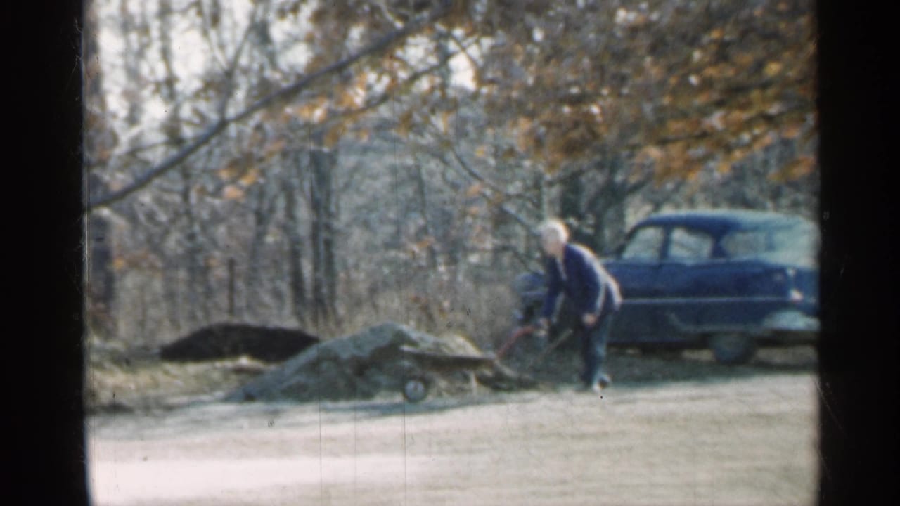 Man bends over with a shovel near dirt piles, wheelbarrow, and parked blue car.