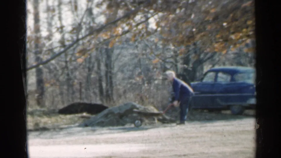 A late-1950s Indiana yard scene shows the man standing over dirt or rocks with a shovel, wheelbarrow, bare trees, and blue sedan visible.