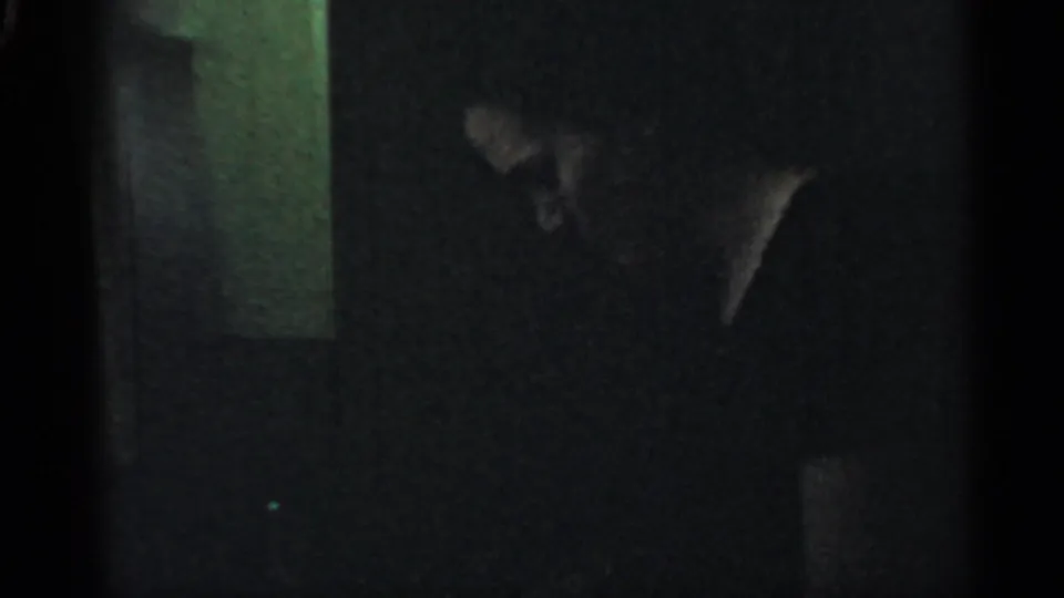 1959 Indiana 8mm frame holding on the dim interior, with a person’s head and shoulder faintly visible near a pale wall edge.