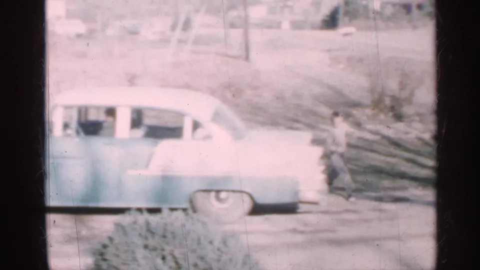 1959 Indiana outdoor frame showing a light-colored rounded sedan on a dirt road while a person moves past carrying long wooden planks.