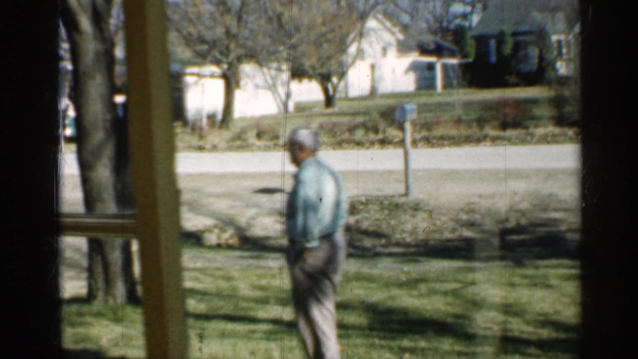 Man in a light shirt walking across a lawn seen through a window frame.