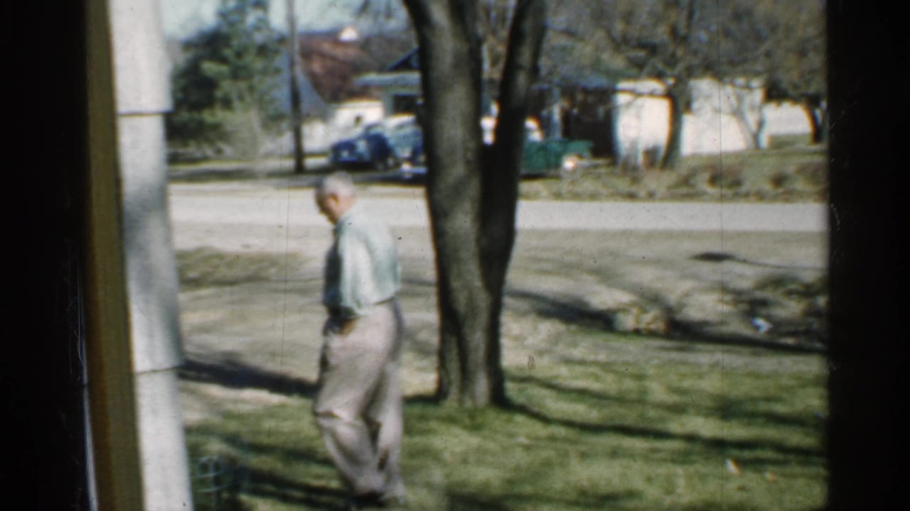 Man standing or walking on grass near trees with houses and a road behind him.