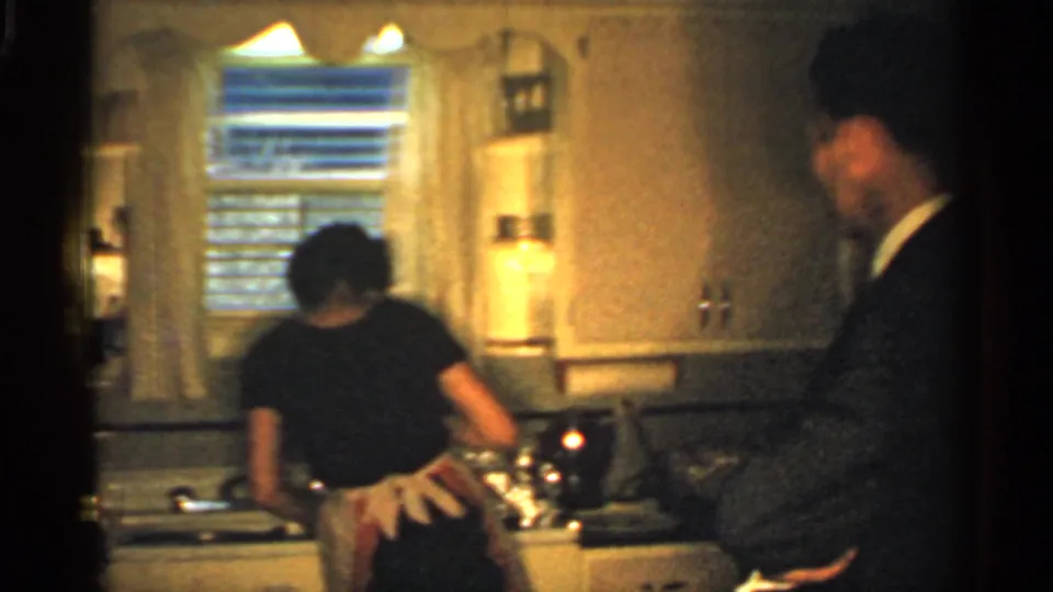 1959 Indiana home kitchen frame showing a woman in an apron at the sink, with a suited man partly visible near the right side.
