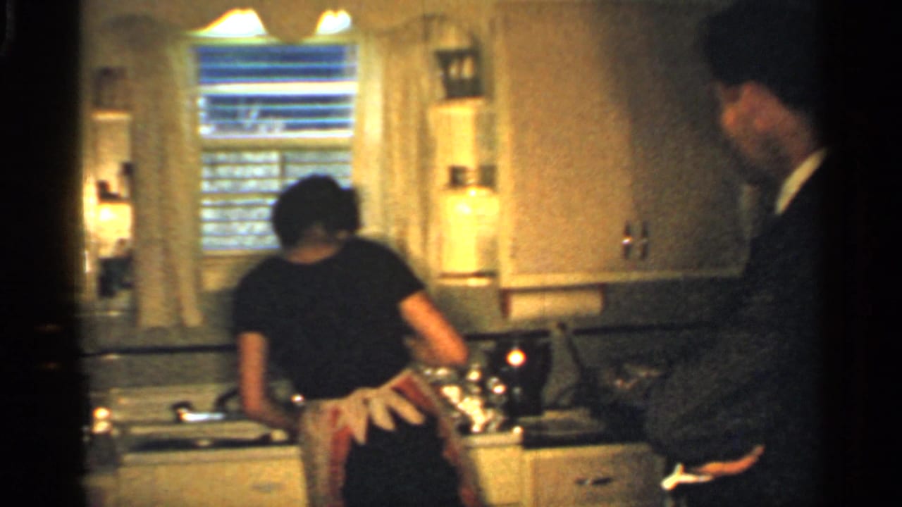 Woman in apron at a kitchen sink as a man in a suit stands nearby.