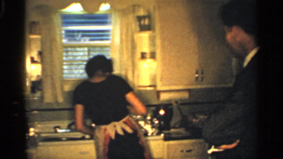 1959 Indiana home-movie frame of a woman working at a kitchen sink while a suited man stands beside the counter.