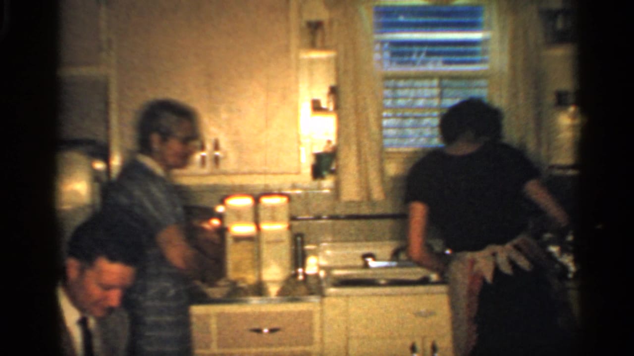 In a 1959 Indiana kitchen, adults stand and sit near the counter, sink, refrigerator, and curtained window.