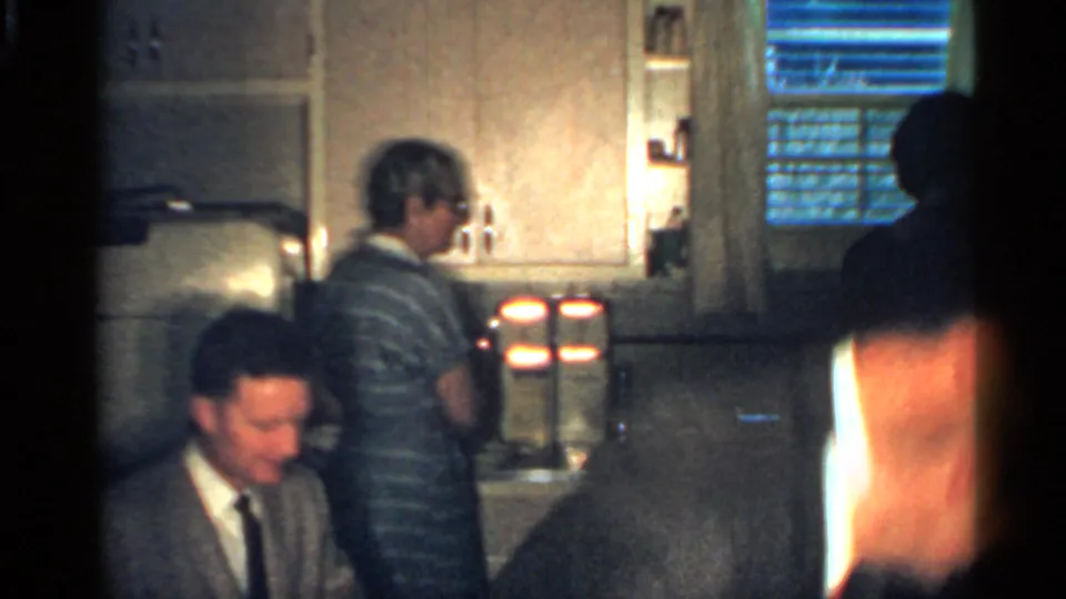 1959 Indiana home-movie frame showing a striped-shirted man by the kitchen counter and a suited man seated in front.
