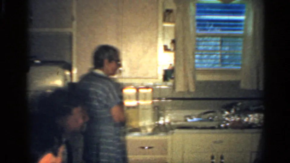 A 1959 Indiana kitchen view with a man at the counter, another adult bending nearby, and sink fixtures under a curtained window.