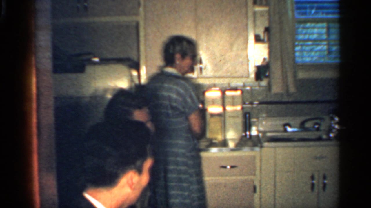 Adults gathered in a small kitchen as a woman in a striped dress works at the counter.