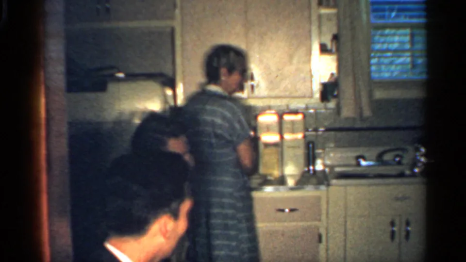 1959 Indiana home-movie frame showing several adults in a compact kitchen while a woman in a striped dress prepares food at the counter.