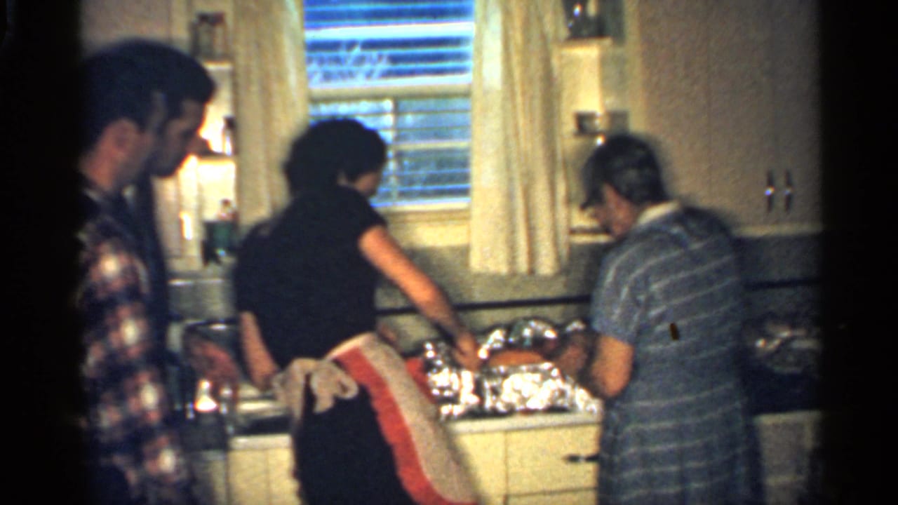 1959 Indiana home-movie frame with two women preparing food in a mid-century kitchen as a man in a plaid shirt watches nearby.