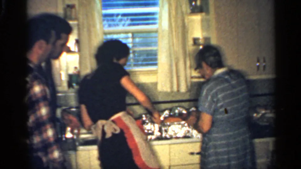 1959 Indiana home-movie frame with two women preparing food in a mid-century kitchen as a man in a plaid shirt watches nearby.