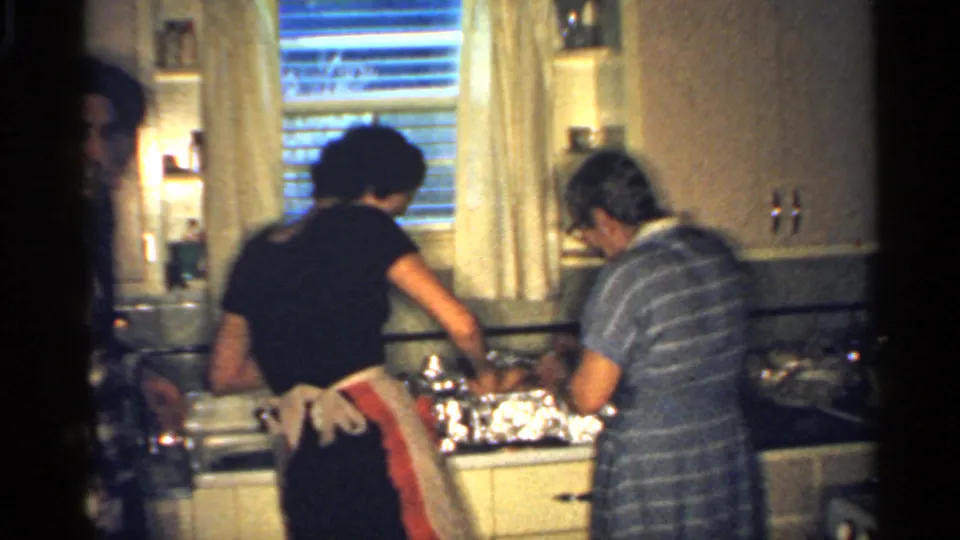 1959 Indiana home-movie frame showing women preparing a meal at a kitchen counter beneath a window with blinds and curtains.