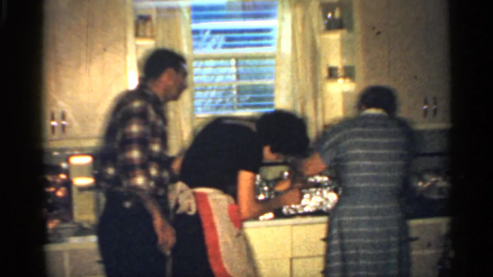 1959 Indiana home-movie frame of a man in a plaid shirt watching as two women lean over food preparation in a private kitchen.