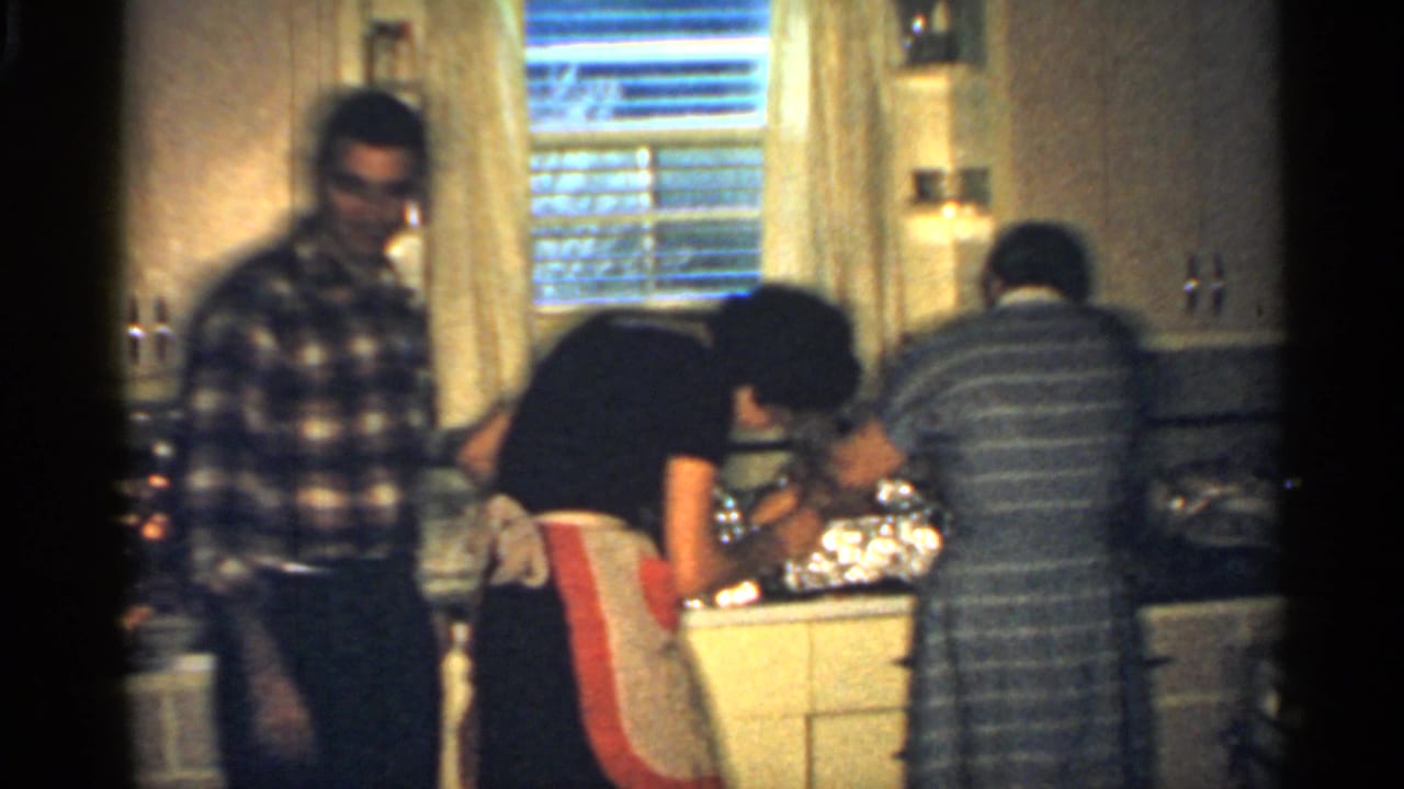 In a 1959 Indiana home-movie frame, three adults work around a mid-century kitchen counter with cabinets, blinds, and foil-covered dishes.