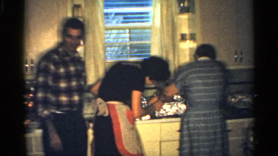 In a 1959 Indiana home-movie frame, three adults work around a mid-century kitchen counter with cabinets, blinds, and foil-covered dishes.