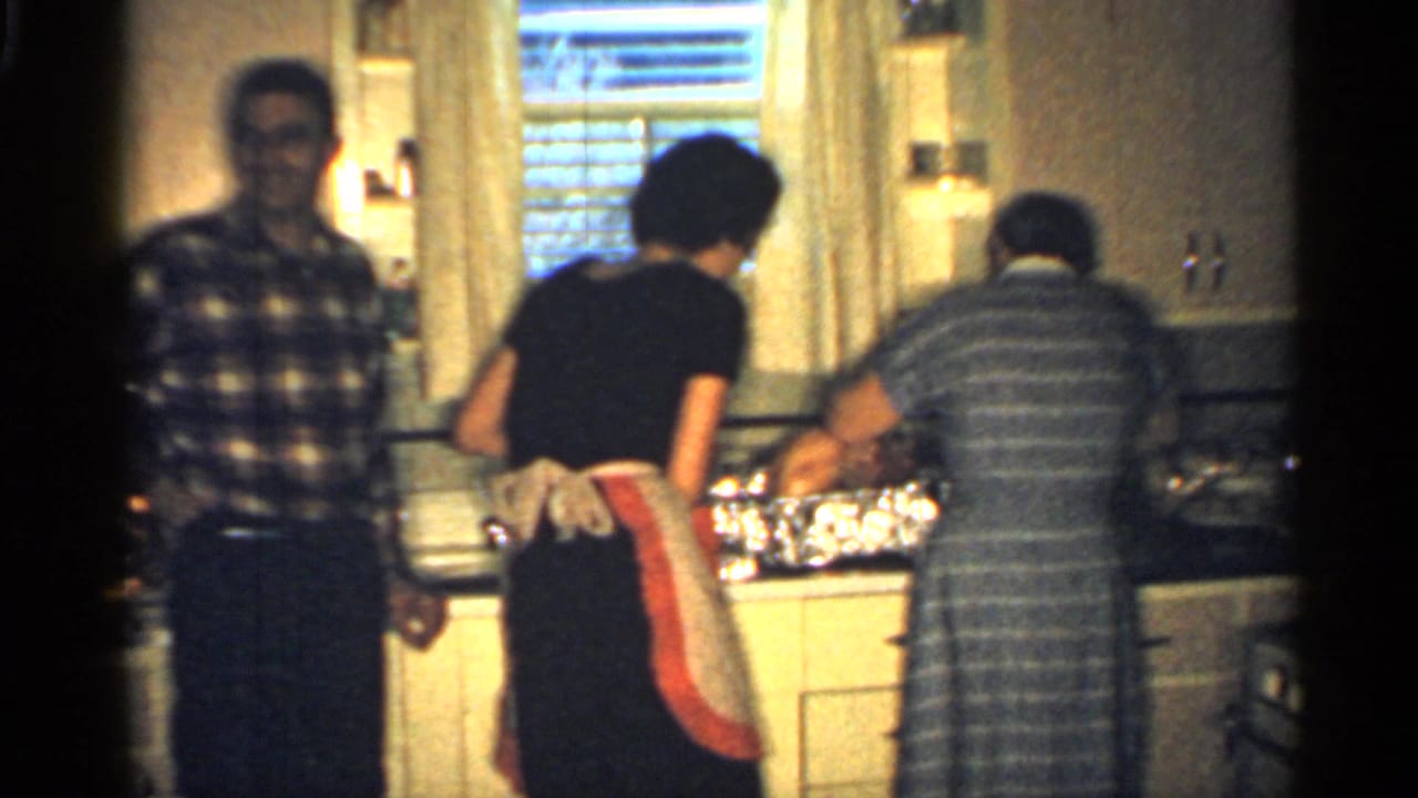 Adults work at a kitchen counter as a man in plaid faces the camera.