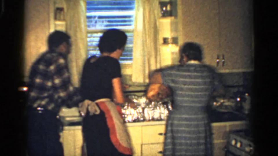Three adults cluster around a compact kitchen work area, with a striped dress, apron, blinds, cabinets, and foil-covered dishes visible.