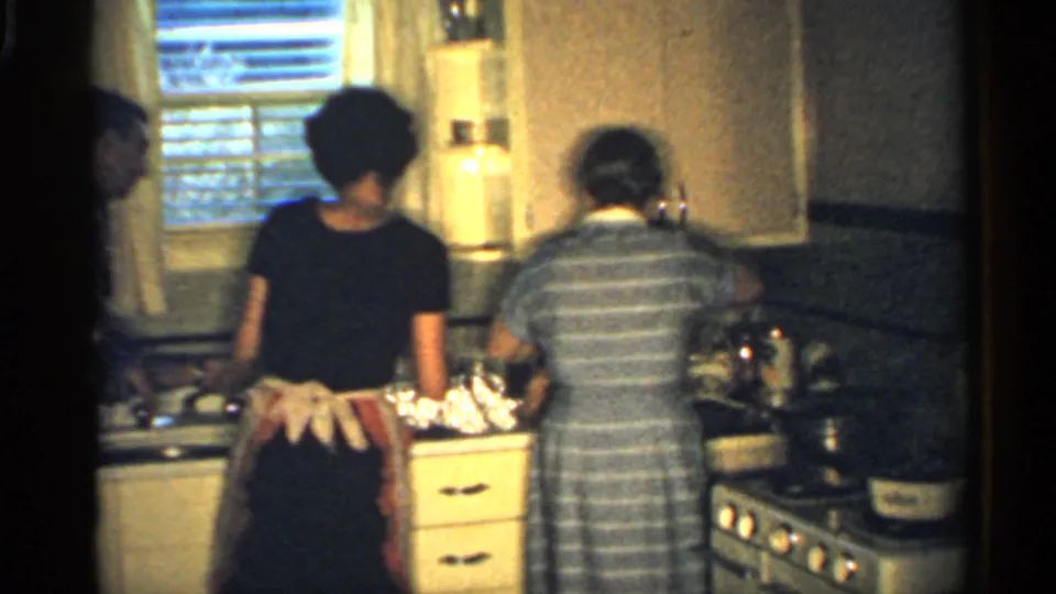 A woman in an apron and another adult in a striped dress stand at a kitchen counter, with blinds, cabinets, cookware, and a stove visible.
