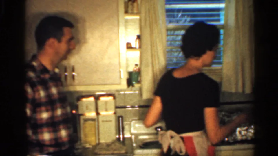 A woman in a black dress and apron faces the kitchen counter while a man in a plaid shirt stands at the side of the frame.