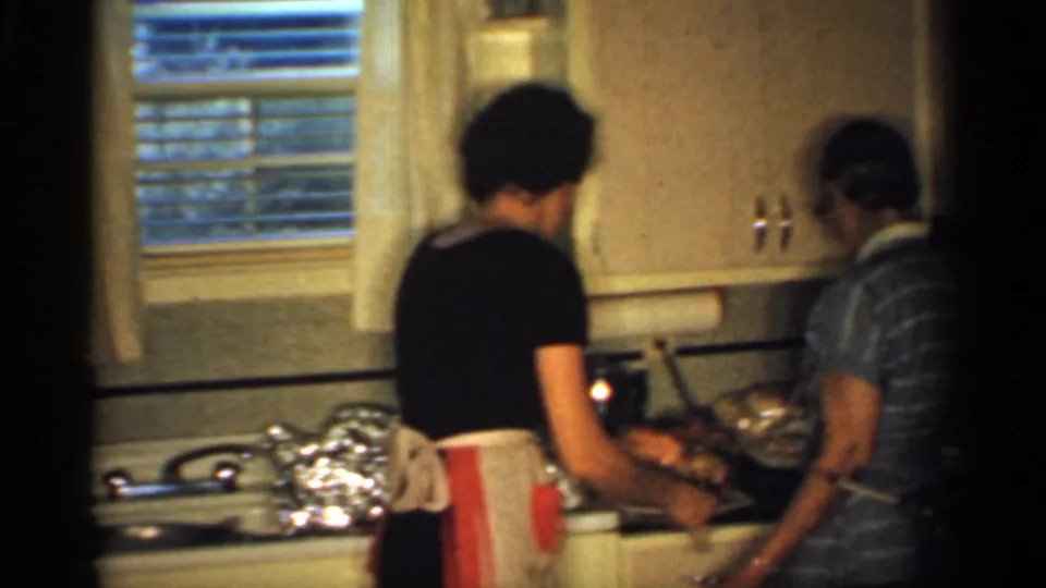 Two women prepare food together at the counter in the 1959 Indiana kitchen, with ham, foil dishes, cabinets, and window visible.
