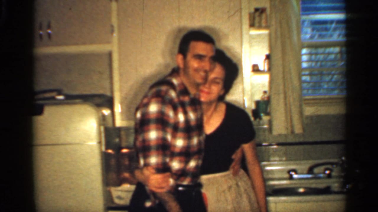 A man in a plaid shirt hugs a woman in a black dress in a kitchen.