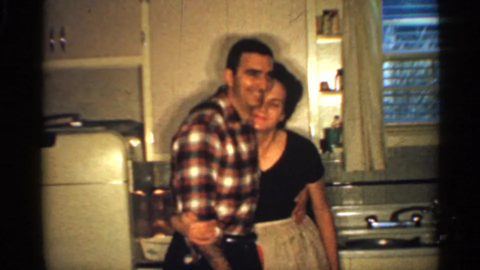 In a 1959 Indiana kitchen, a man in a plaid shirt embraces a woman in a black dress as they face the camera.
