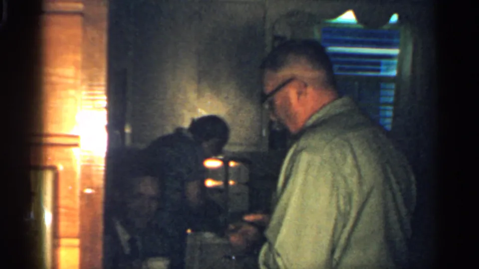 In the kitchen interior, an older man stands near the counter while another person remains partly obscured in shadow.