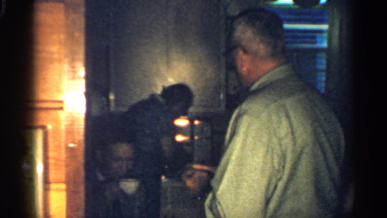 1959 Indiana home-movie frame showing men gathered in a dim private interior with warm light at the edge.