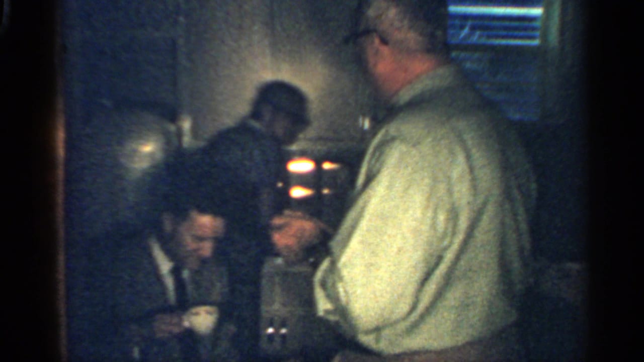 Dim 1959 Indiana interior with an older man in a light shirt near seated men and cups on or near a table.