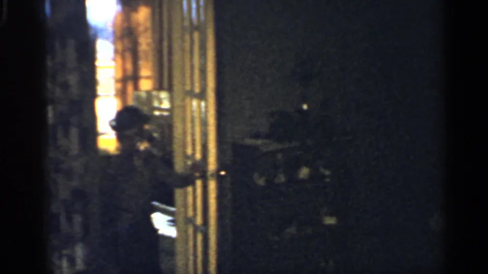 1959 Indiana home-movie frame showing a woman in a light dress opening or standing at a doorway in a dim private room.