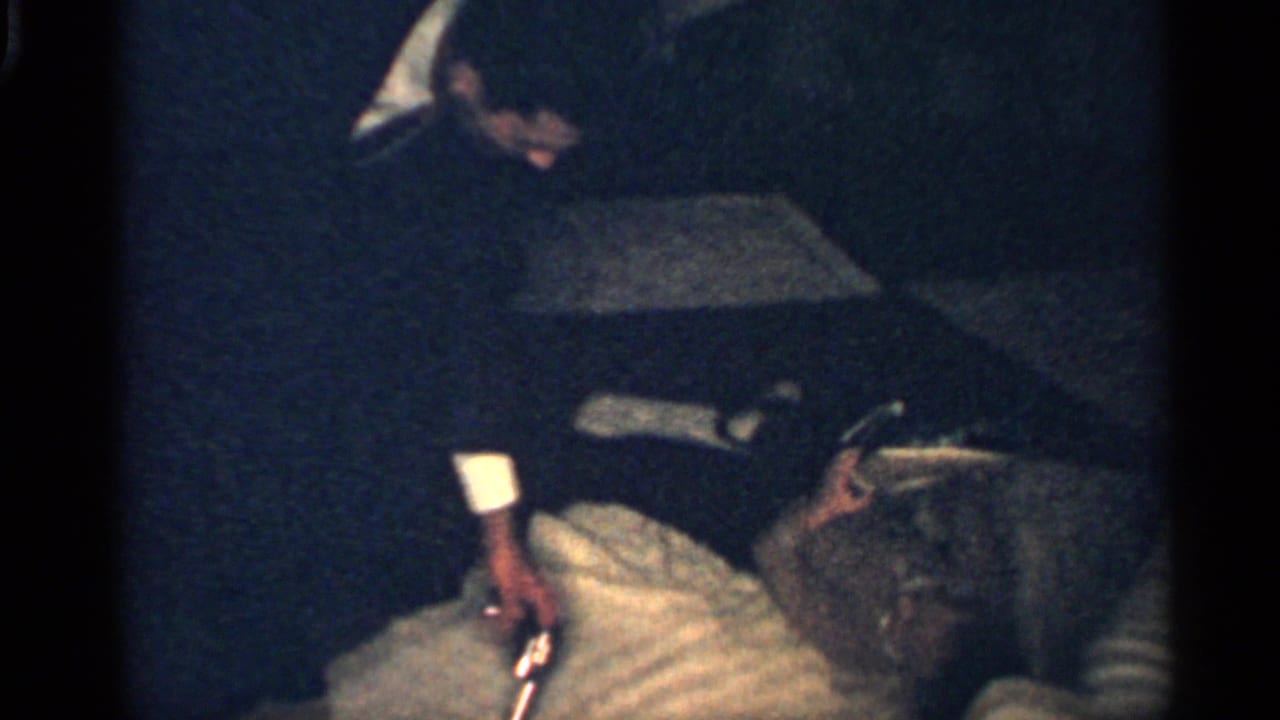 In a dark 1959 Indiana interior, the boy remains on the bed while adult figures stand close by and a small object is visible near him.